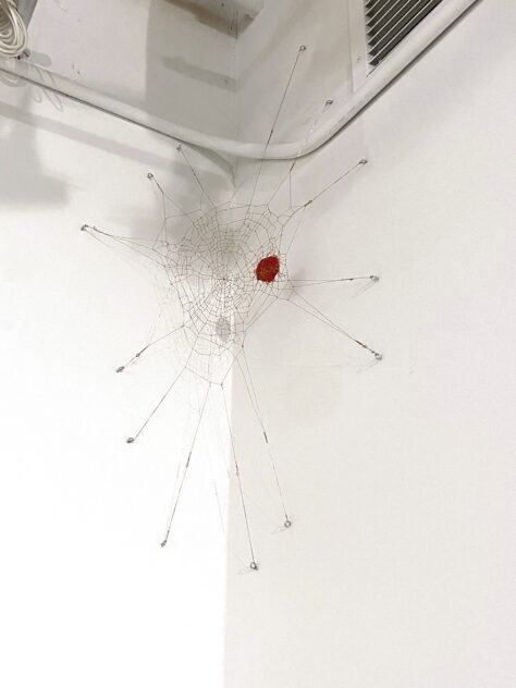 UUUUUU (The Last Laugh) by Bradly Brown — copper-wire spiderweb holding a red clown nose, strung below a ceiling pipe in a white gallery