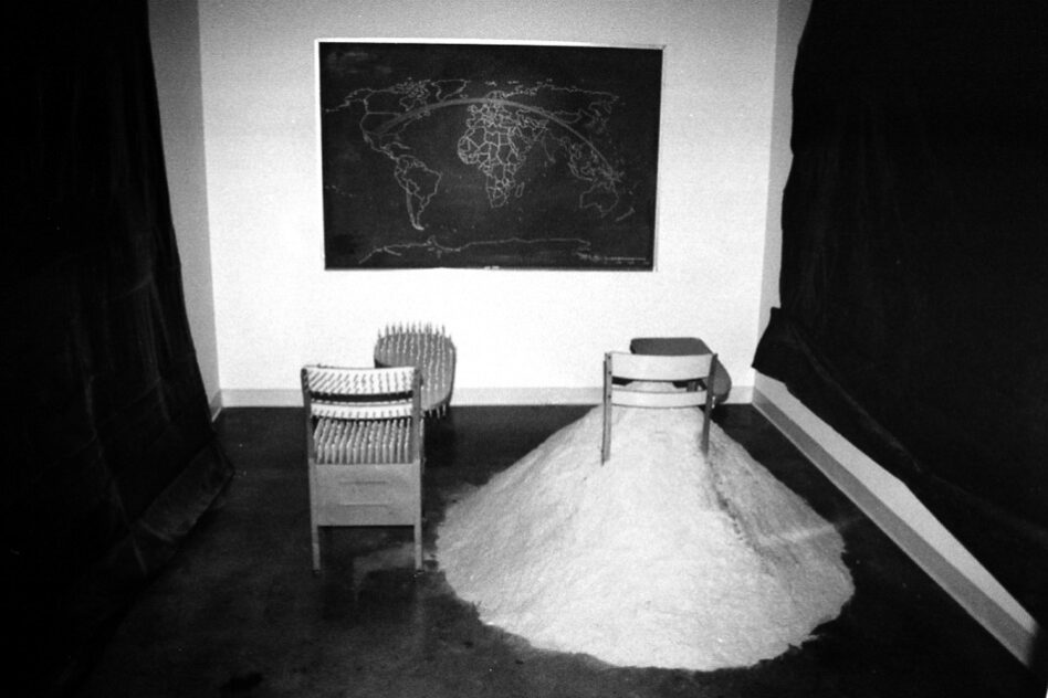 15,540 Miles, 2002 — artwork by artist Bradly Brown. Black and white view of the two school desks, one buried in sawdust, beneath the chalkboard map