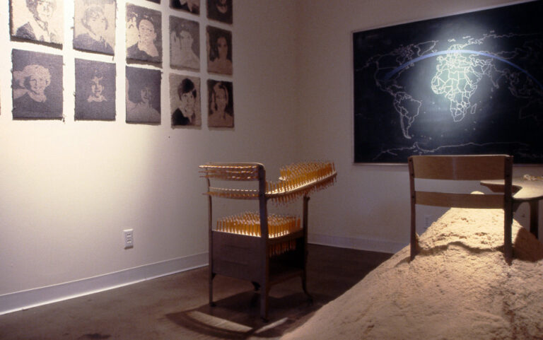 15,540 Miles, 2002 — artwork by artist Bradly Brown. Installation view at Conduit Gallery: two school desks in front of a chalkboard world map, with carpet portraits on the side wall