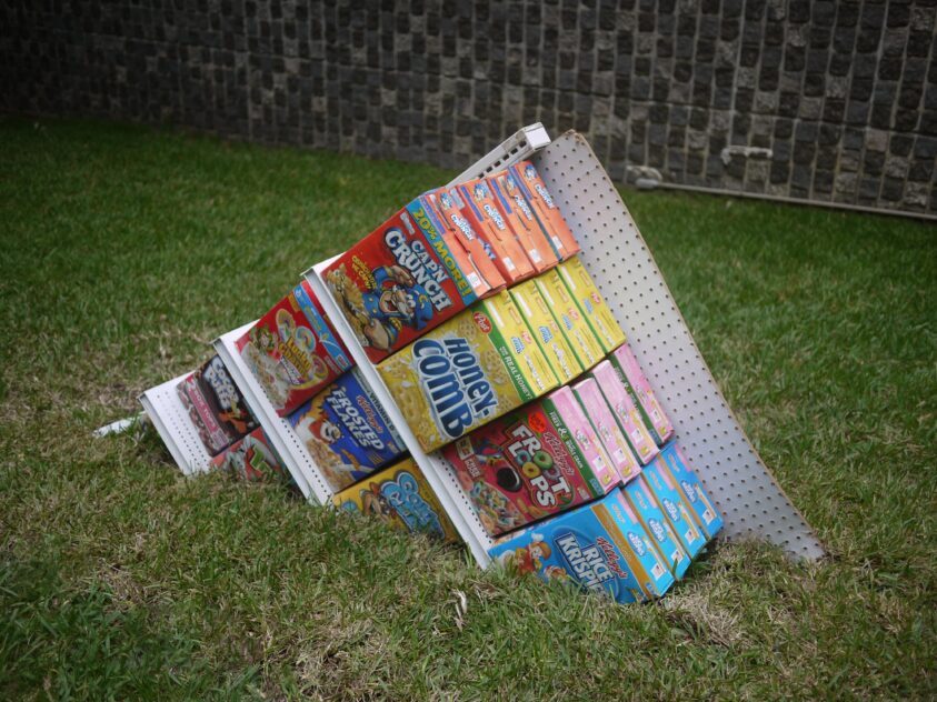 Partially Buried Cereal Aisle, 2013 — artwork by artist Bradly Brown. Gondola shelving stocked with cereal boxes, tilted and half-sunk into a grass lawn