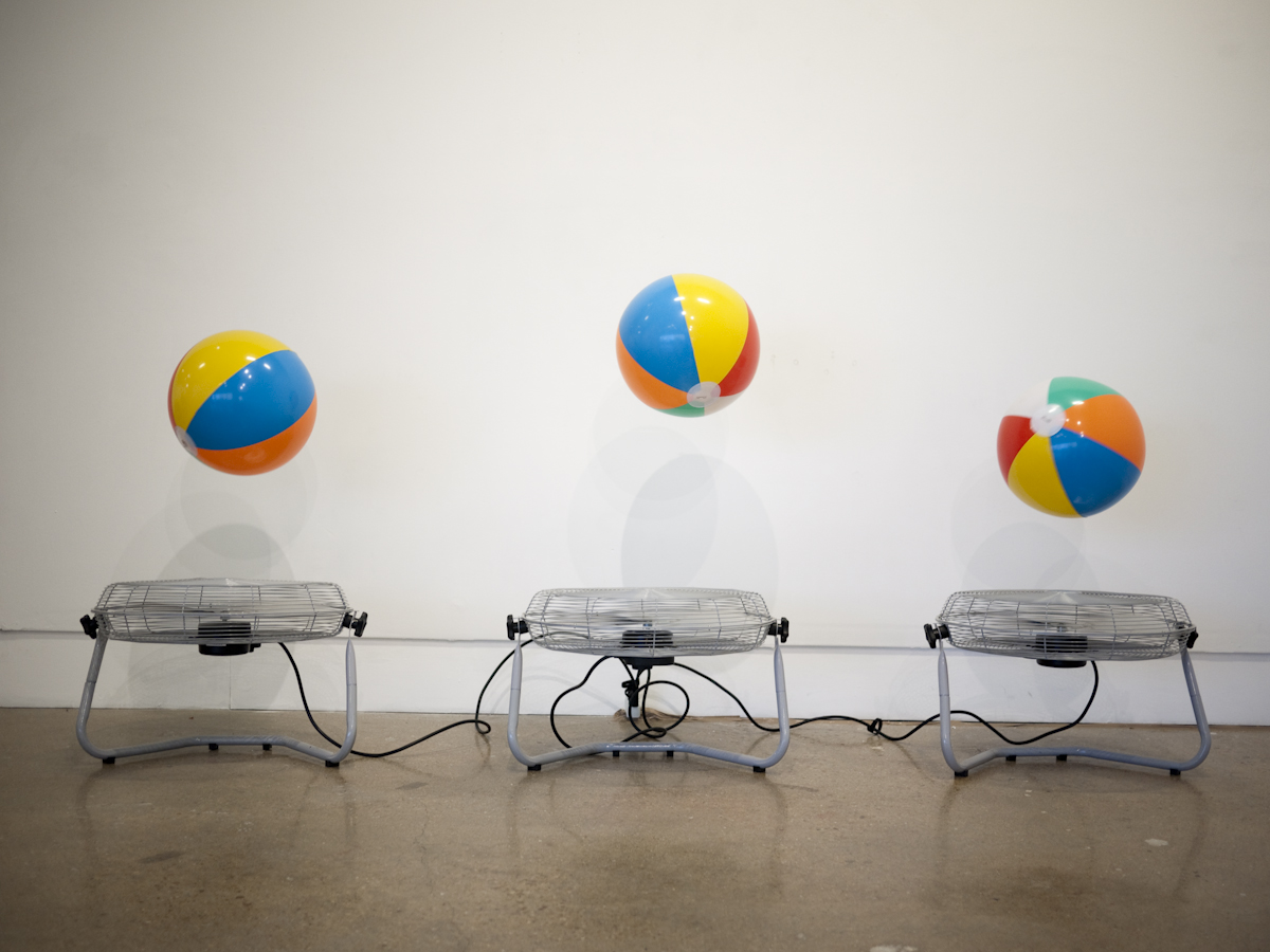 Attention Retention, 2012 — artwork by artist Bradly Brown. Three chrome floor fans in a row, each levitating a striped beach ball in the gallery