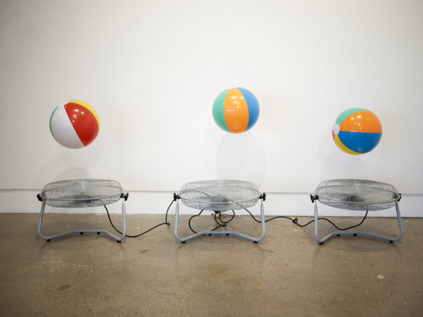 Three beach balls hovering above three floor fans against a white gallery wall. Attention Retention by Bradly Brown